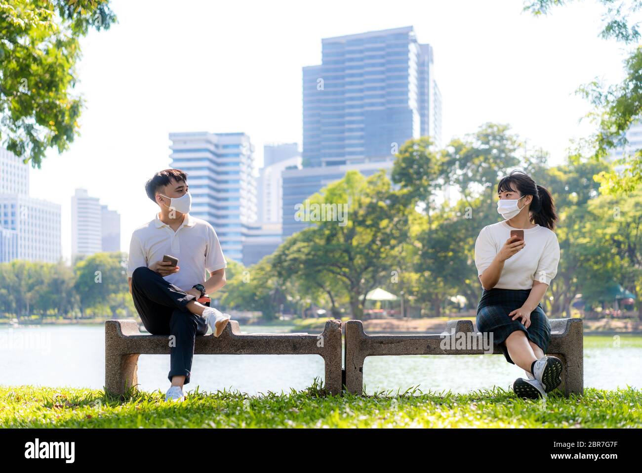 Deux jeunes hommes et femmes asiatiques qui discutent dans un smartphone et portent un masque assis à une distance de 6 pieds protègent contre les virus COVID-19 pour les dis sociaux Banque D'Images
