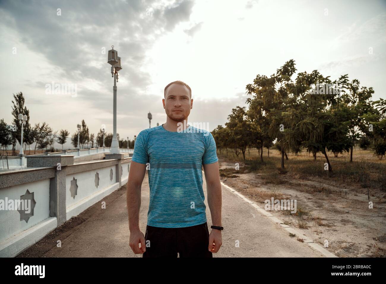 Portrait of Caucasian guy dans un tee-shirt bleu et un short noir qui s'entraîne et s'exécute sur la piste asphaltée pendant le coucher du soleil Banque D'Images