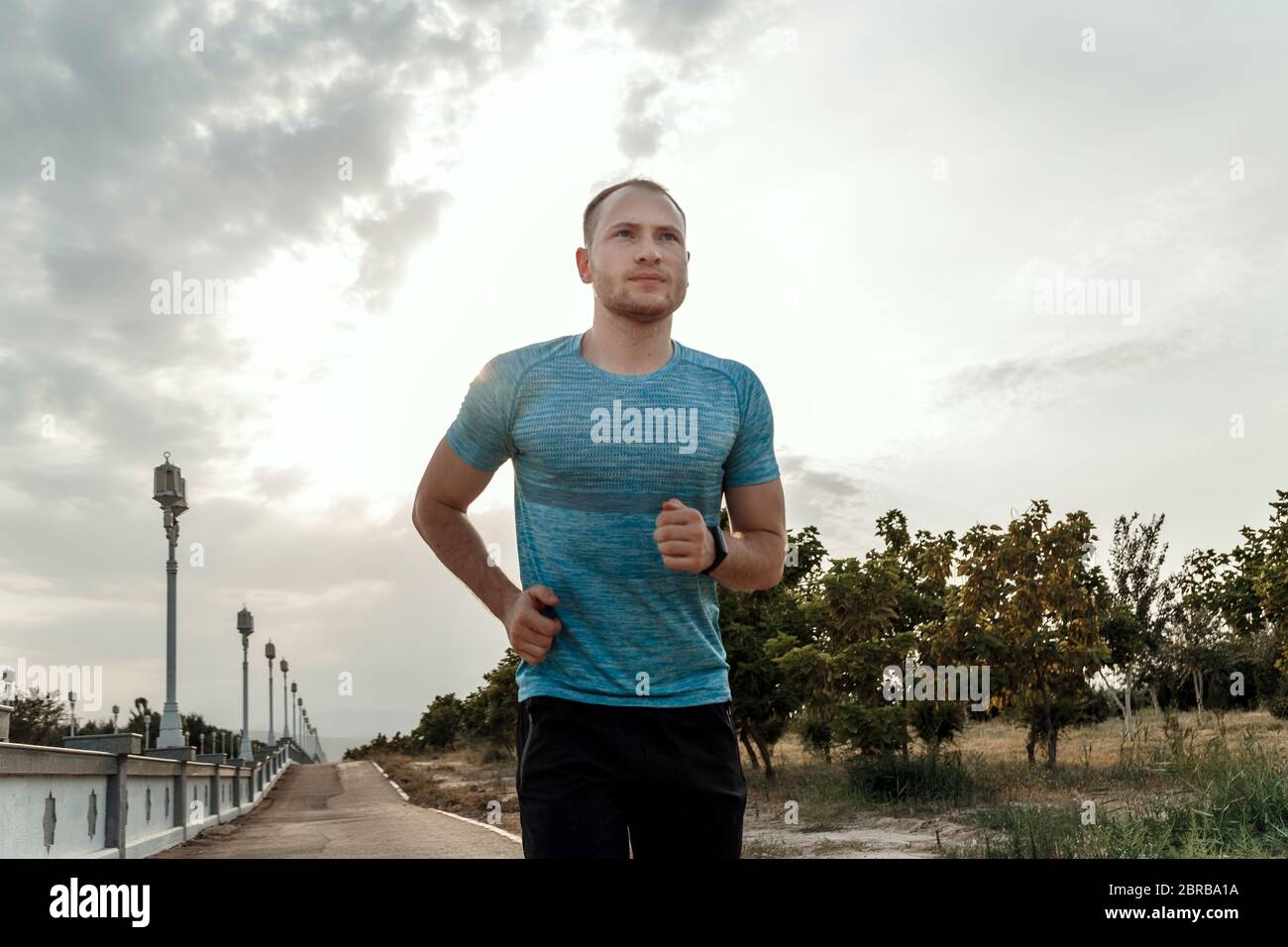 Portrait of Caucasian guy dans un tee-shirt bleu et un short noir qui s'entraîne et s'exécute sur la piste asphaltée pendant le coucher du soleil Banque D'Images