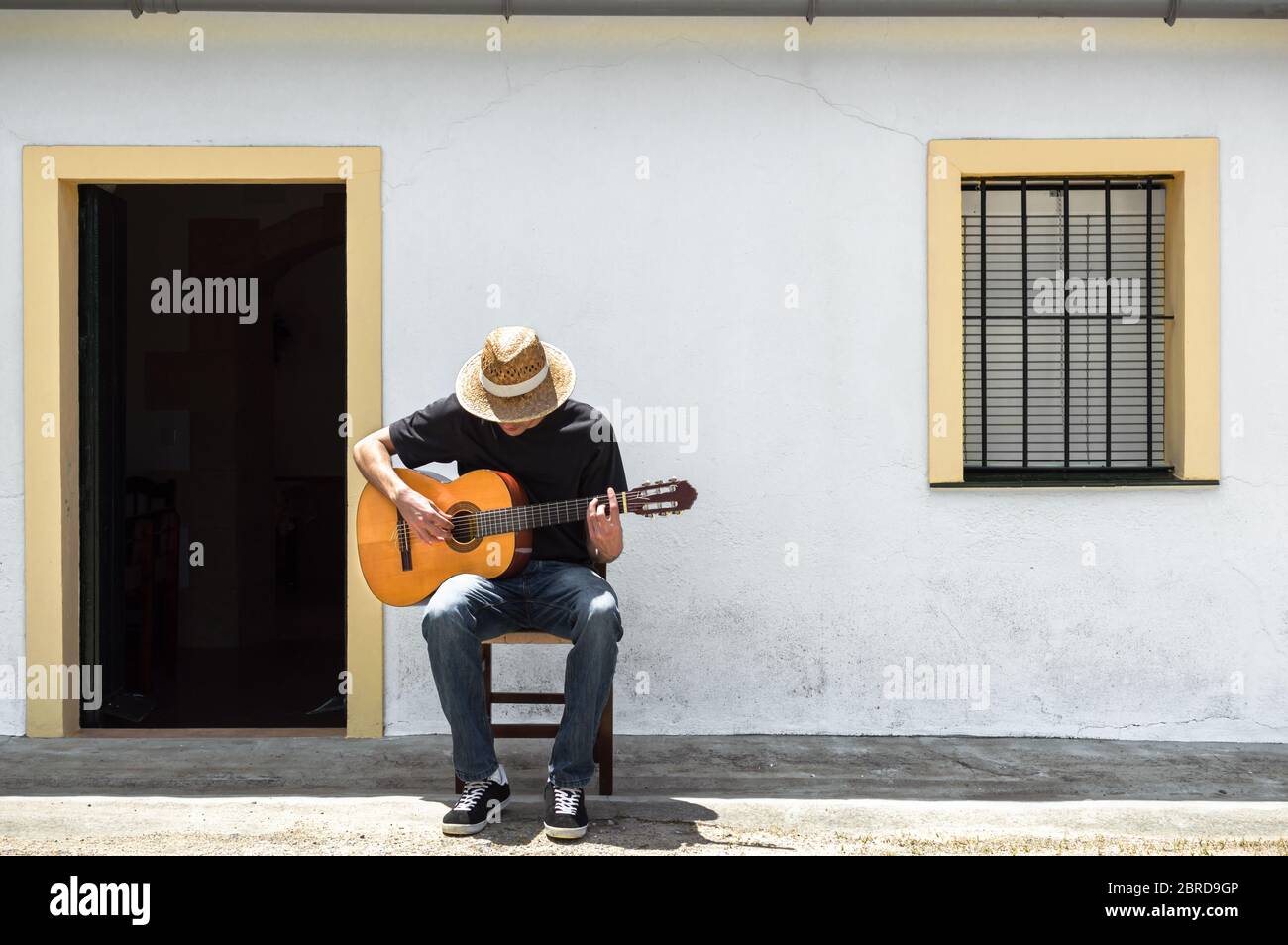 Jeune homme avec chapeau de paille jouant de la guitare classique devant la façade de sa maison blanche à la campagne par une journée ensoleillée. Mode de vie rural. Banque D'Images