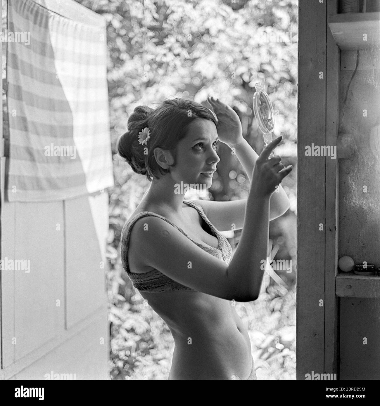 jeune jolie femme debout dans la porte de la maison en utilisant le miroir de main le jour chaud ensoleillé portant bikini années 1960 hongrie Banque D'Images