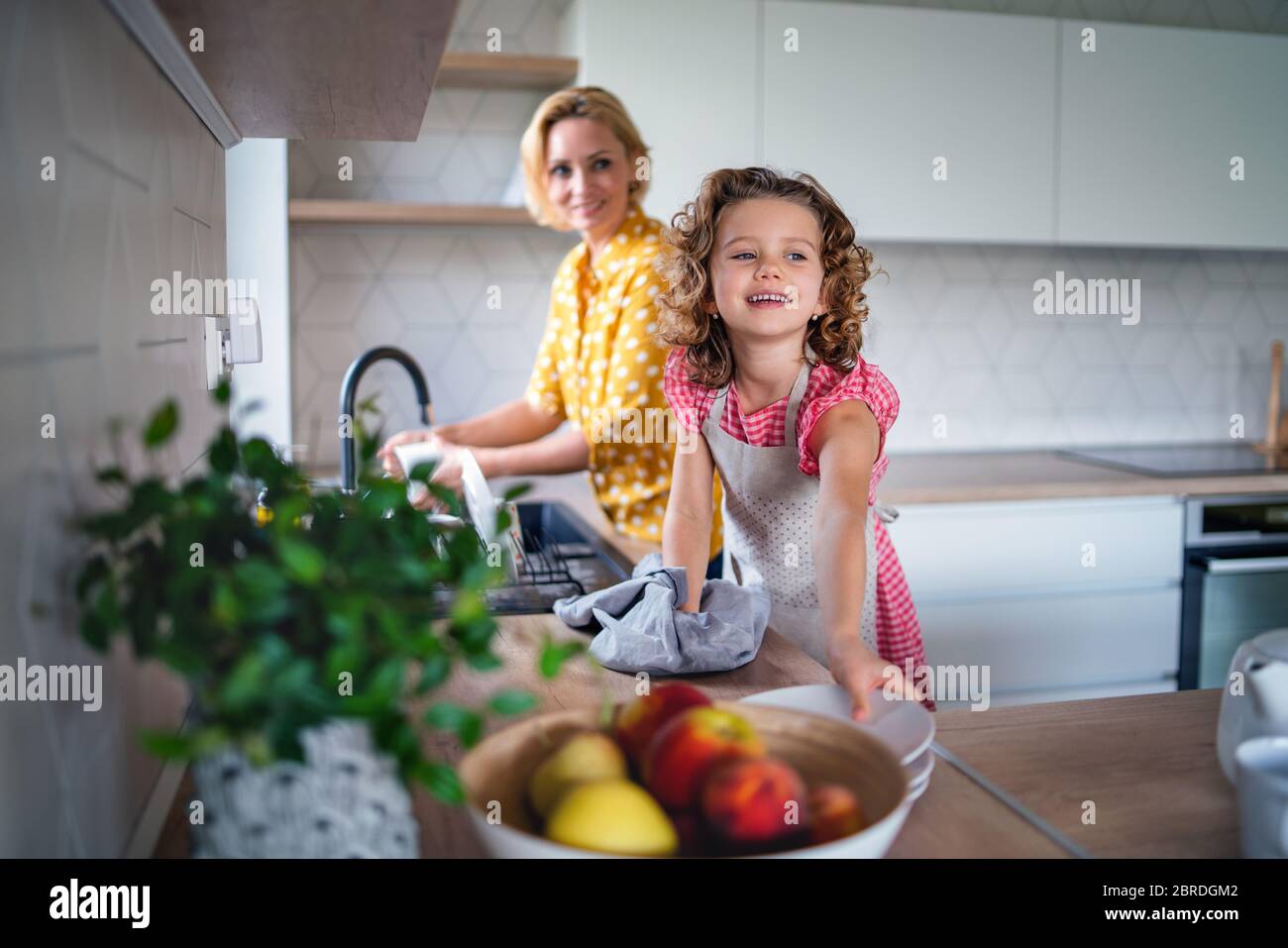 Une petite fille mignonne avec une mère à l'intérieur dans la cuisine à la maison, laver la vaisselle. Banque D'Images