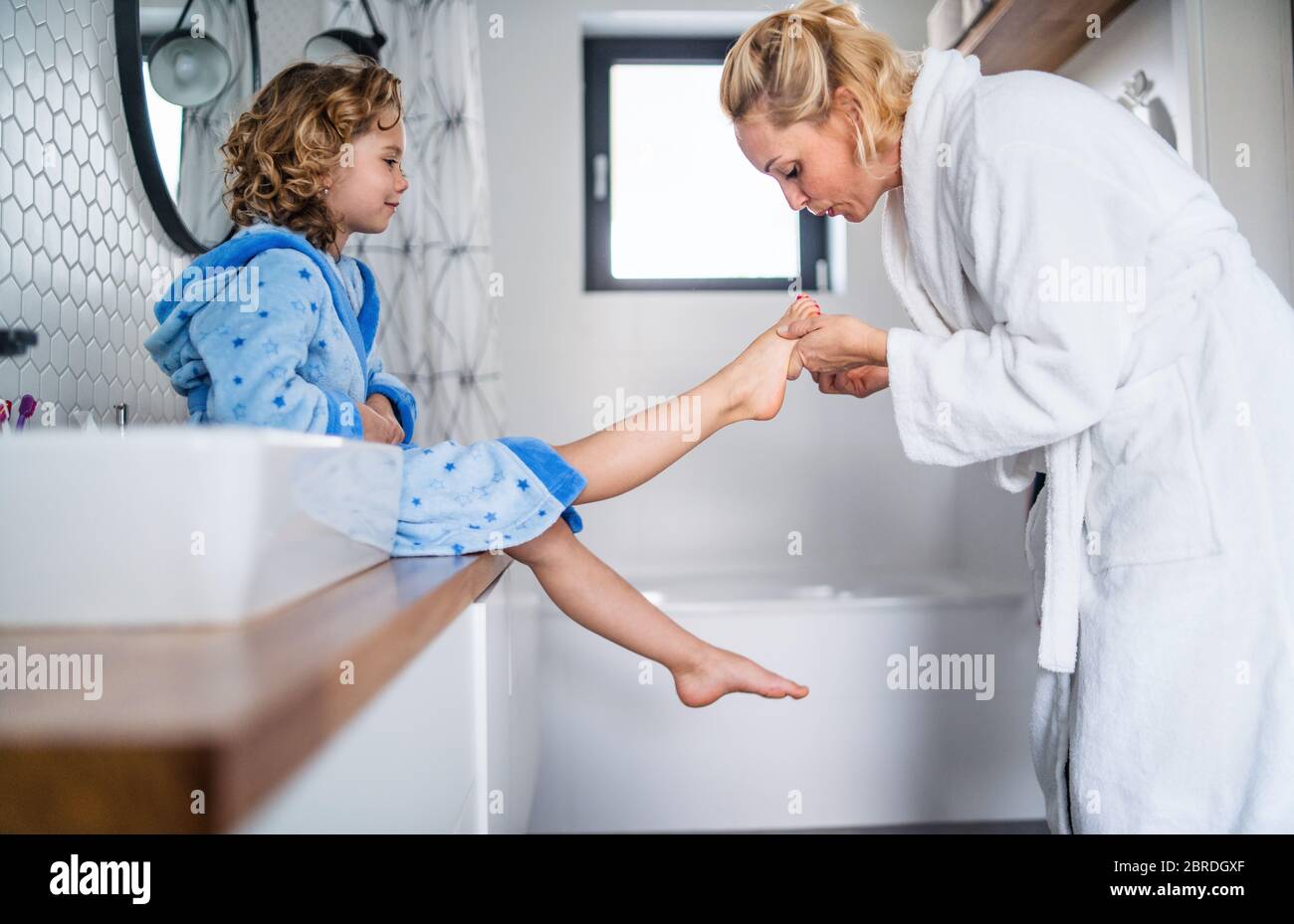 Une petite fille mignonne avec une mère dans la salle de bains à l'intérieur à la maison, peindre des ongles. Banque D'Images