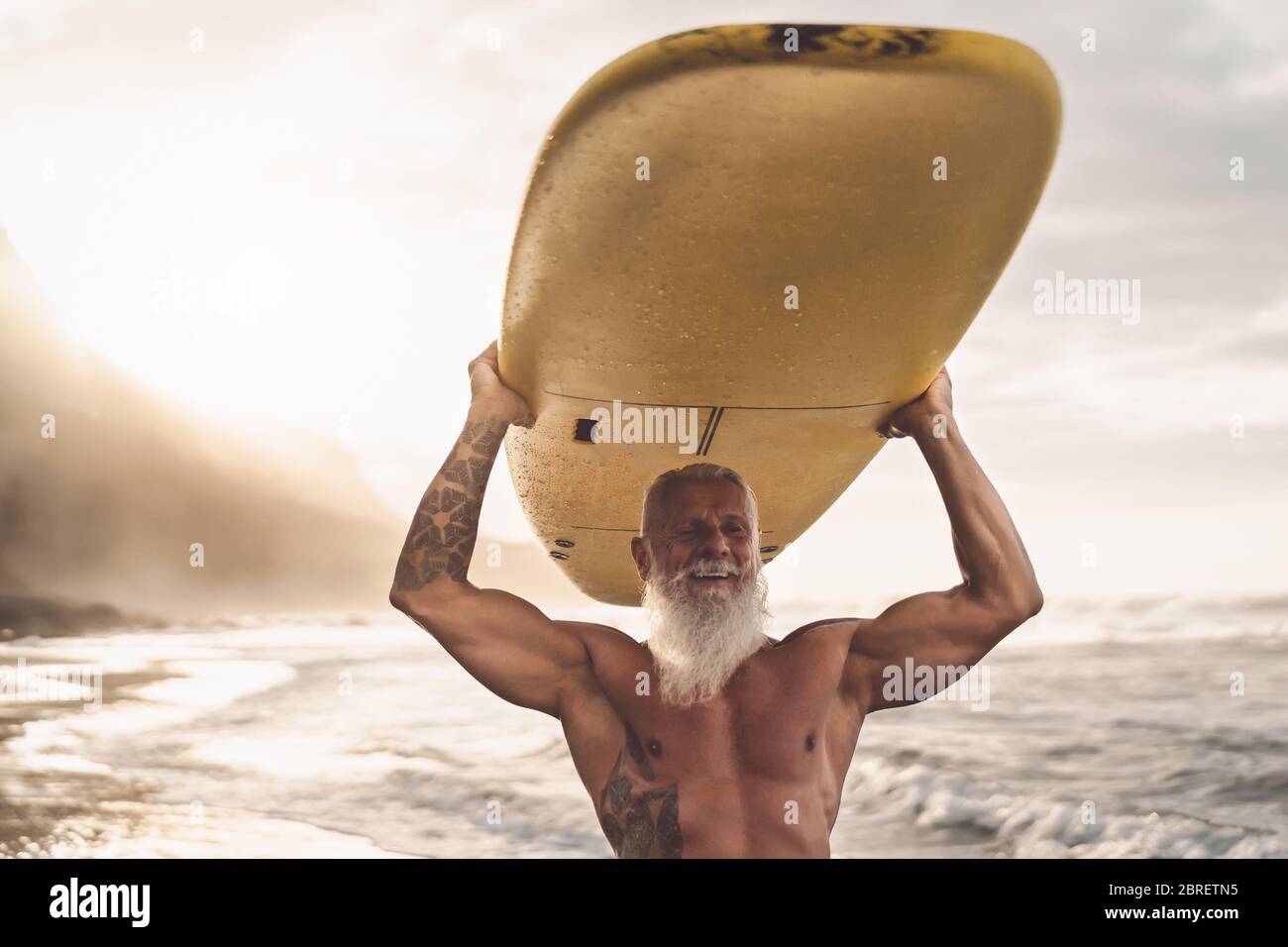 Bonne tenue pour les personnes âgées qui s'amusent à surfer au coucher du soleil - Sporty barbu homme s'entraîner avec planche de surf sur la plage Banque D'Images
