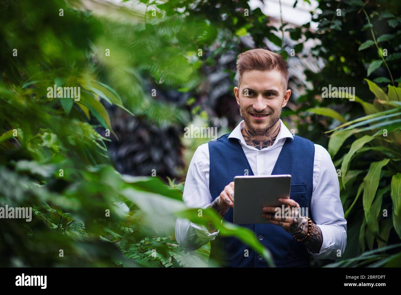 Jeune homme avec une tablette debout dans le jardin botanique, travaillant. Banque D'Images