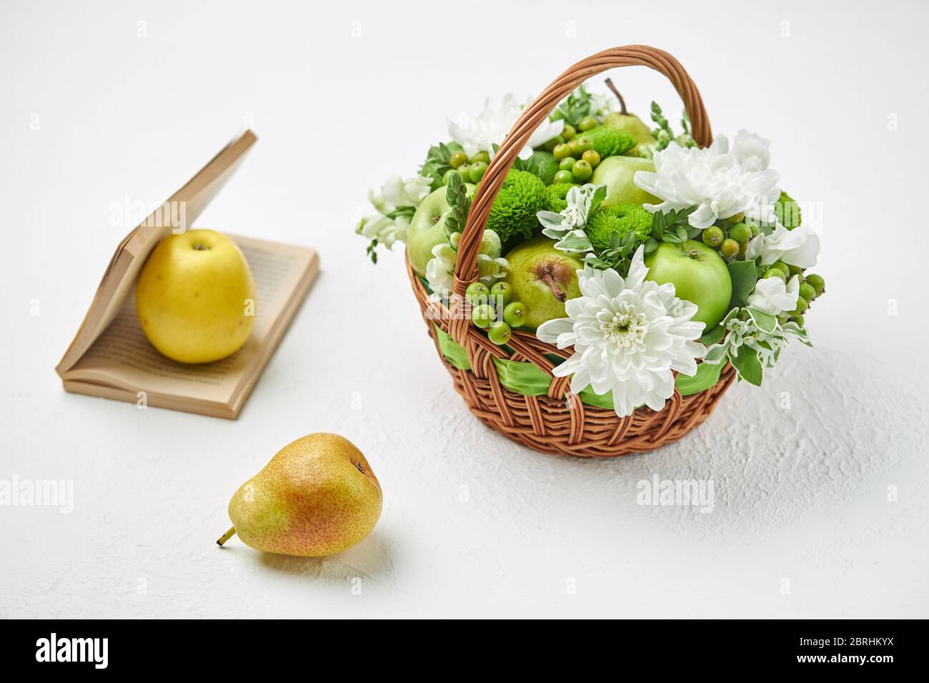 Encore la vie avec un panier en osier rempli de fruits, de fleurs blanches et un livre ouvert. Banque D'Images