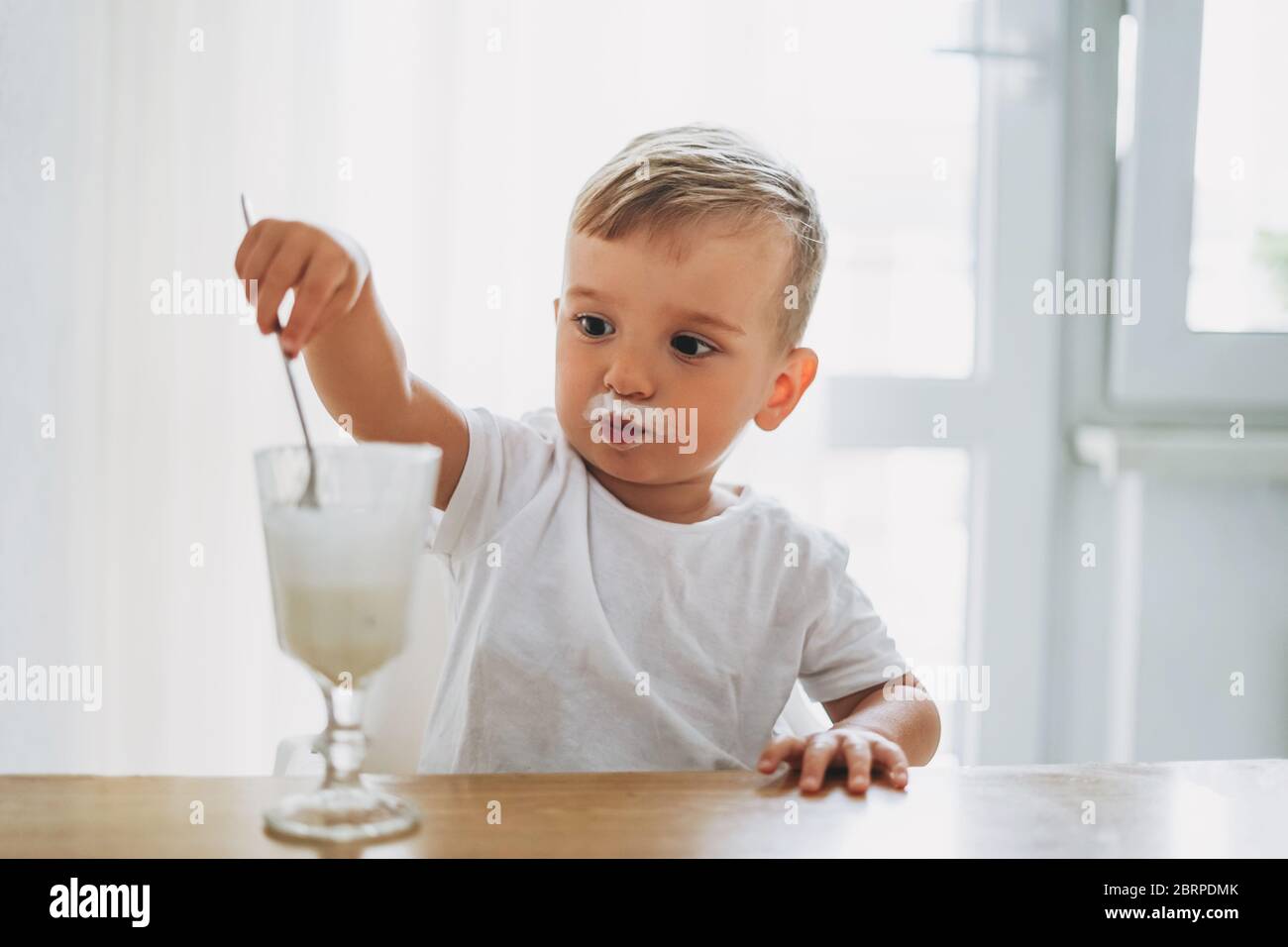 Charmant petit garçon concentré manger du yaourt en verre dans la cuisine lumineuse à la maison Banque D'Images