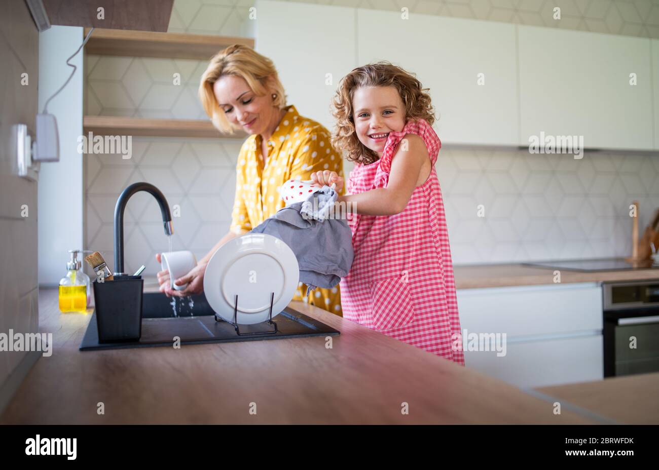 Une petite fille mignonne avec une mère à l'intérieur dans la cuisine à la maison, laver la vaisselle. Banque D'Images