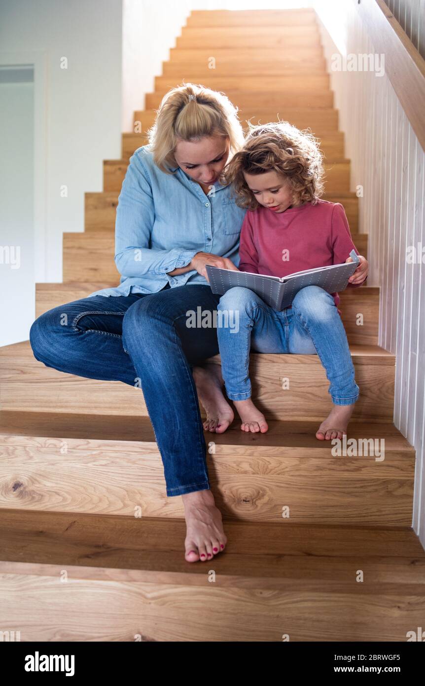 Une petite fille mignonne avec une mère à l'intérieur à la maison, lisant sur l'escalier. Banque D'Images