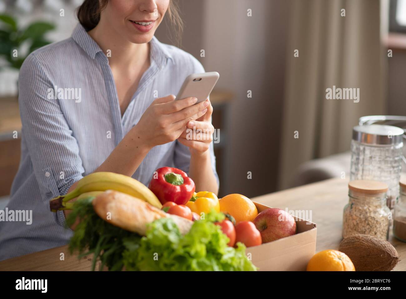 Fille en casque d'intérieur gris assise dans la cuisine avec un smartphone et en tapant un message Banque D'Images