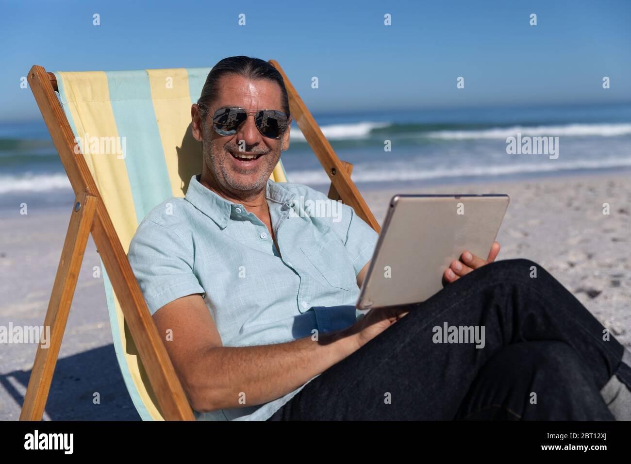 Homme caucasien senior assis sur une chaise longue et utilisant une tablette numérique à la plage Banque D'Images