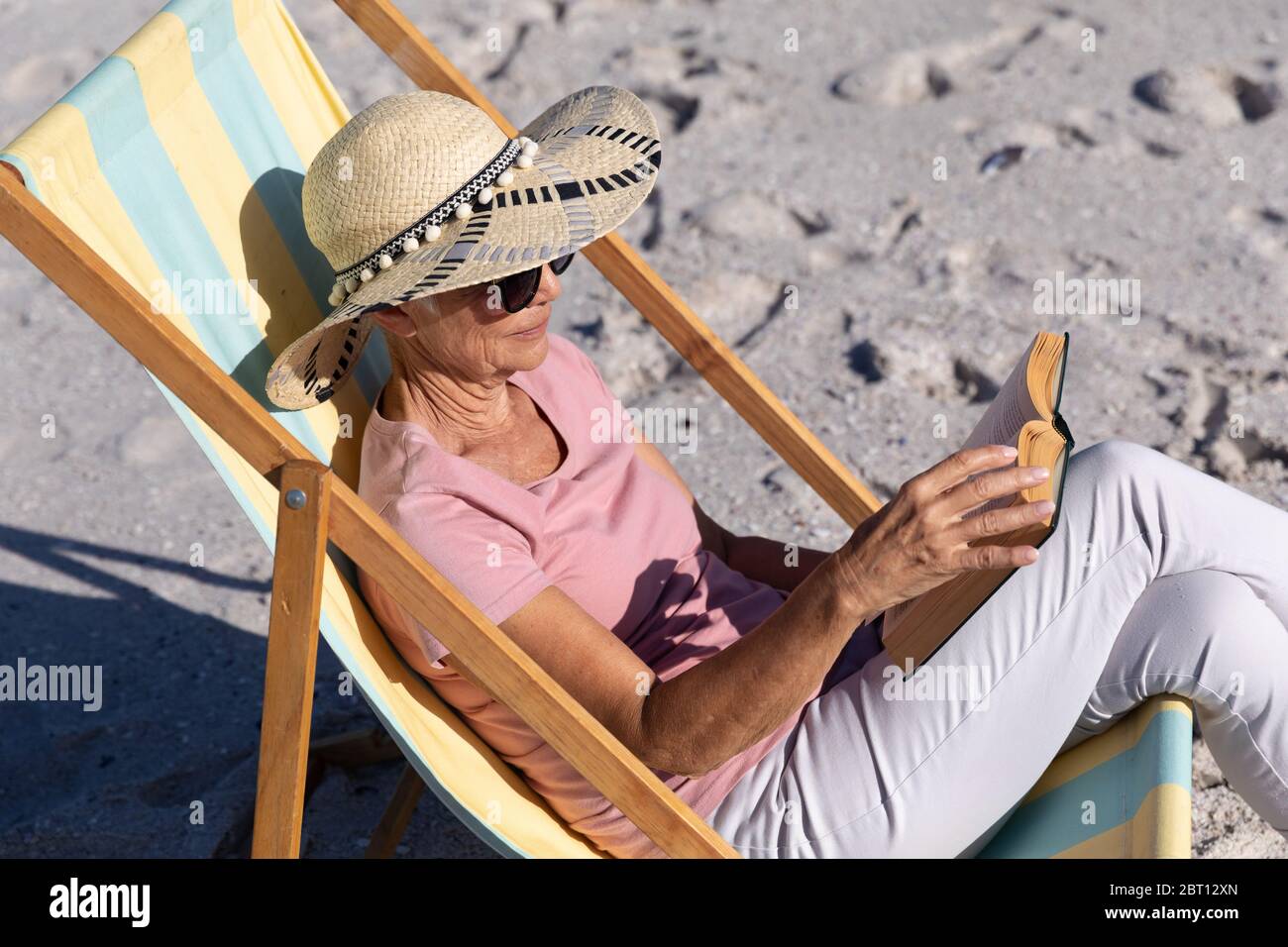 Femme de race blanche assise sur une chaise longue et lisant à la plage. Banque D'Images