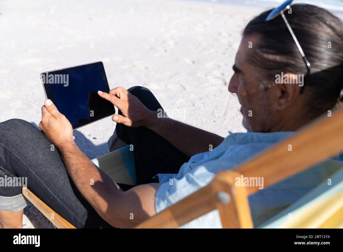 Homme caucasien senior assis sur une chaise longue et utilisant une tablette numérique à la plage Banque D'Images