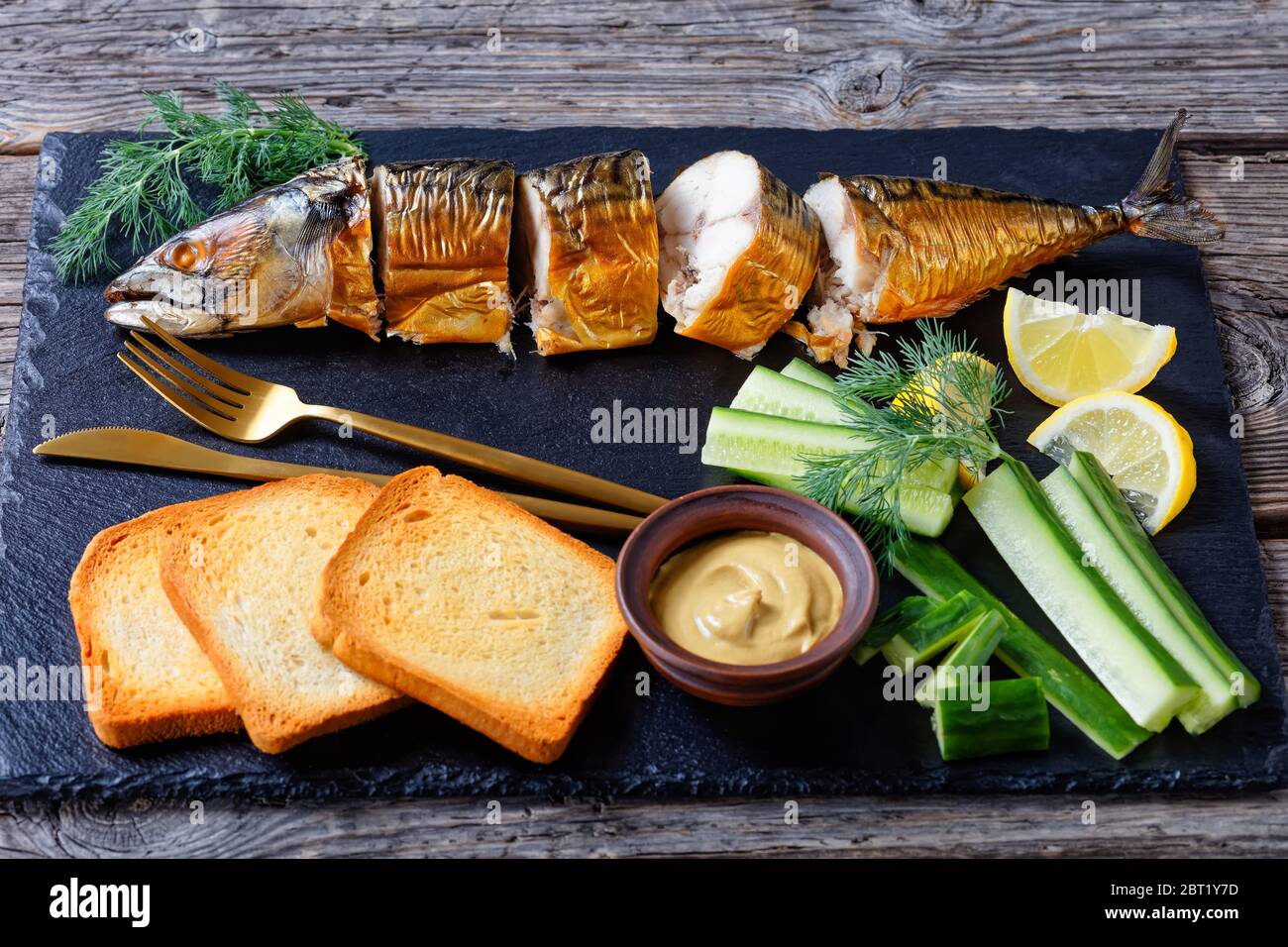 Poisson gras : maquereau fumé chaud servi sur un tableau noir avec moutarde de dijon, concombre frais, toasts, quartiers de citron et couverts sur un dorsard en bois Banque D'Images