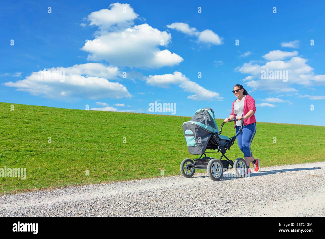 Jeune mère souriante poussant poussette bébé. Herbe verte et ciel bleu avec fond de nuages Banque D'Images