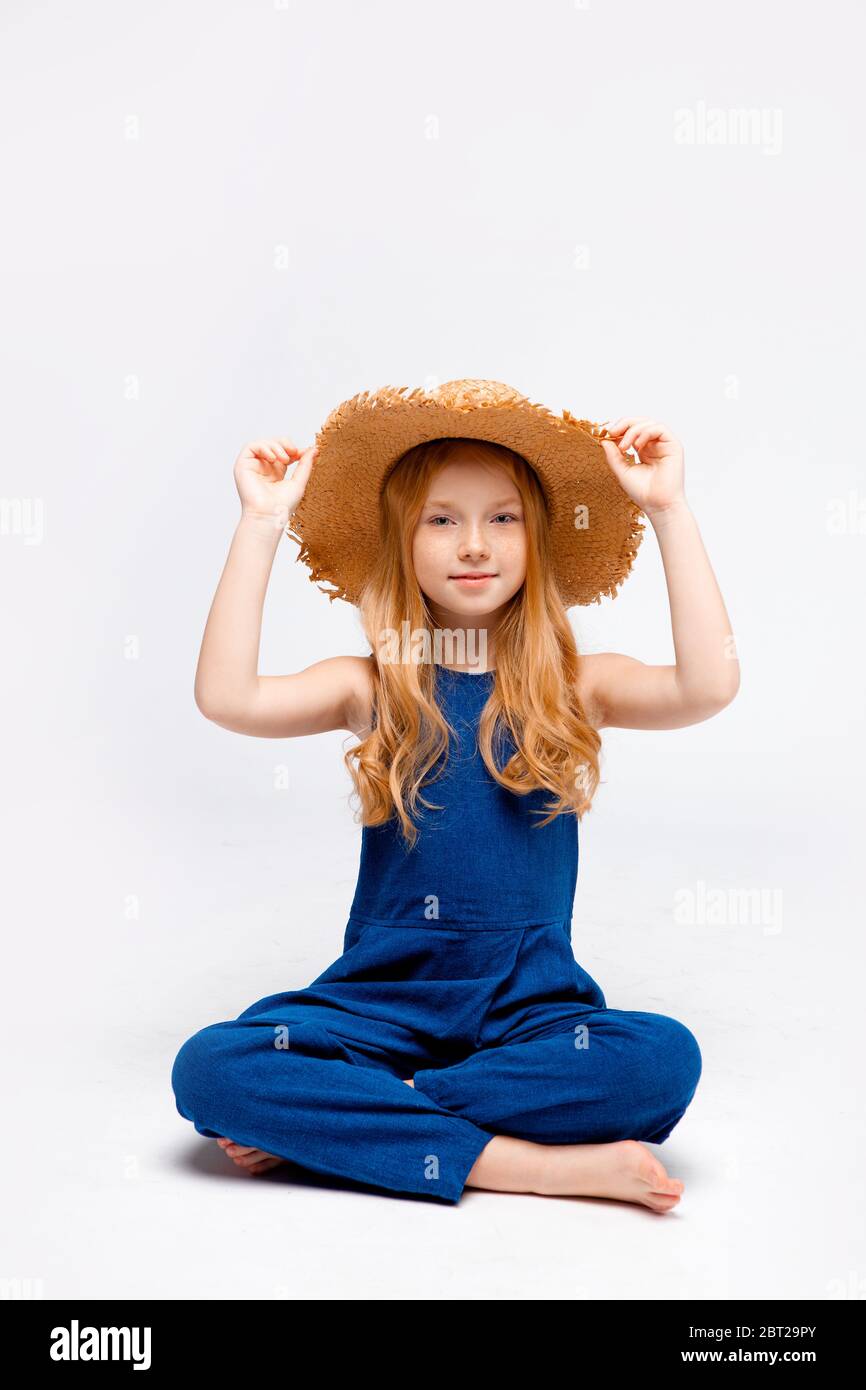 fille à cheveux rouges assise dans une combinaison bleue et un chapeau de paille, jambes croisées Banque D'Images