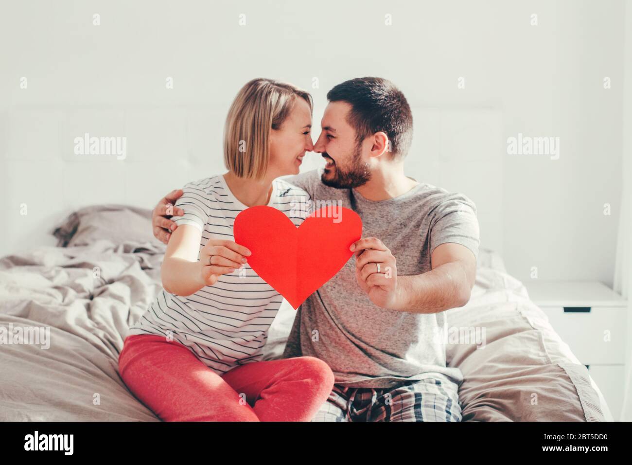 Heureux Mariage Fort Heureux Couple Drole Dans L Amour Tenant Le Cœur De Papier Rouge Homme De Famille Heterosexuel Et Femme Assis Sur Le Lit Dans La Chambre A La Maison Embrassant Photo