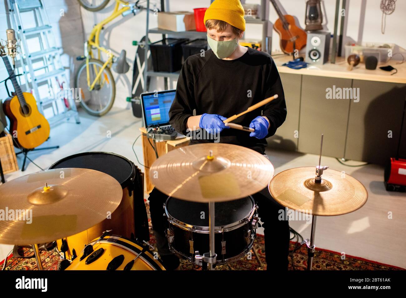 Jeune musicien en gants et masque de protection assis par le tambour dans le garage ou le studio pendant la quarantaine et frapper des cymbales et des tambours Banque D'Images