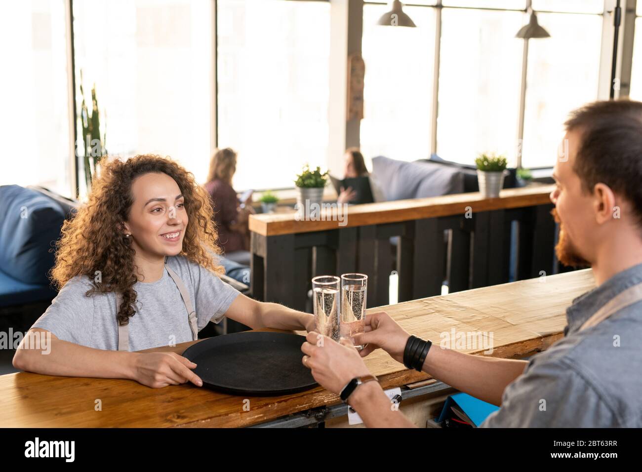 Bonne serveuse jeune avec plateau regardant son collègue en comptoir mettant deux verres d'eau pour les clients de café ou de restaurant Banque D'Images