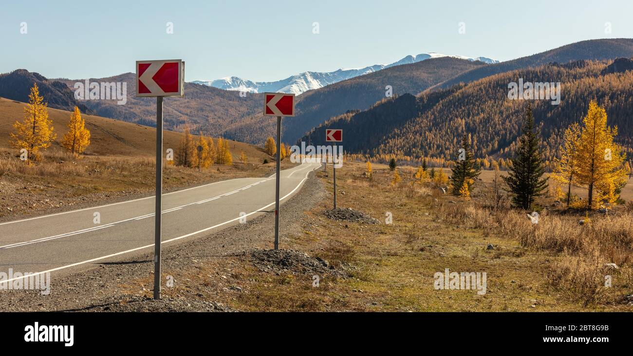 Vue panoramique sur une autoroute dans les montagnes de l'Altaï. Panneaux rouges et blancs sur le trottoir avec flèches orientées vers la gauche. Pics enneigés en arrière-plan Banque D'Images