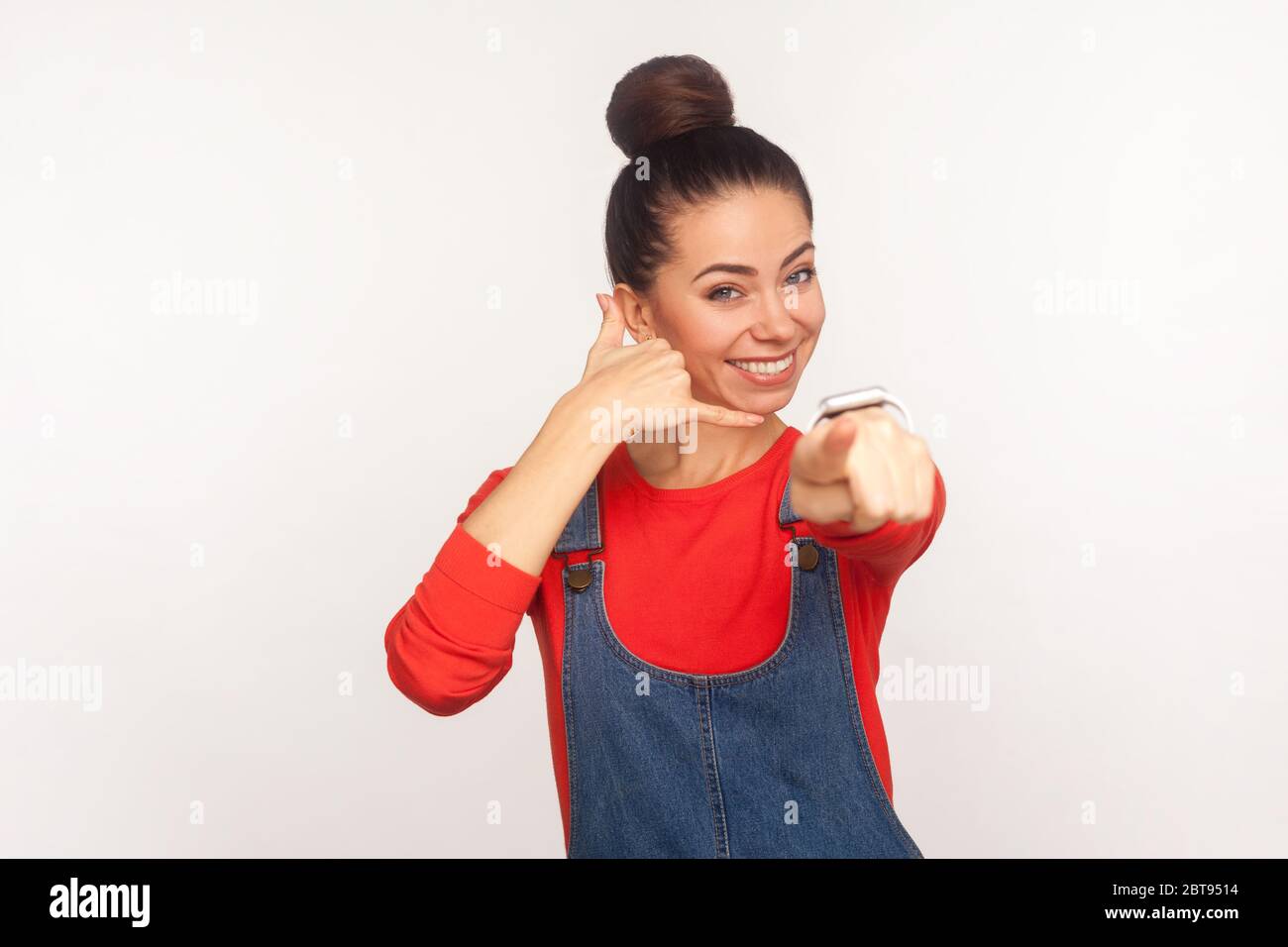 Contactez-nous par téléphone ! Portrait de gai élégant jolie fille avec un pain de cheveux dans des combinaisons en denim montrant appelez-moi geste et pointant vers l'appareil photo. indoo Banque D'Images