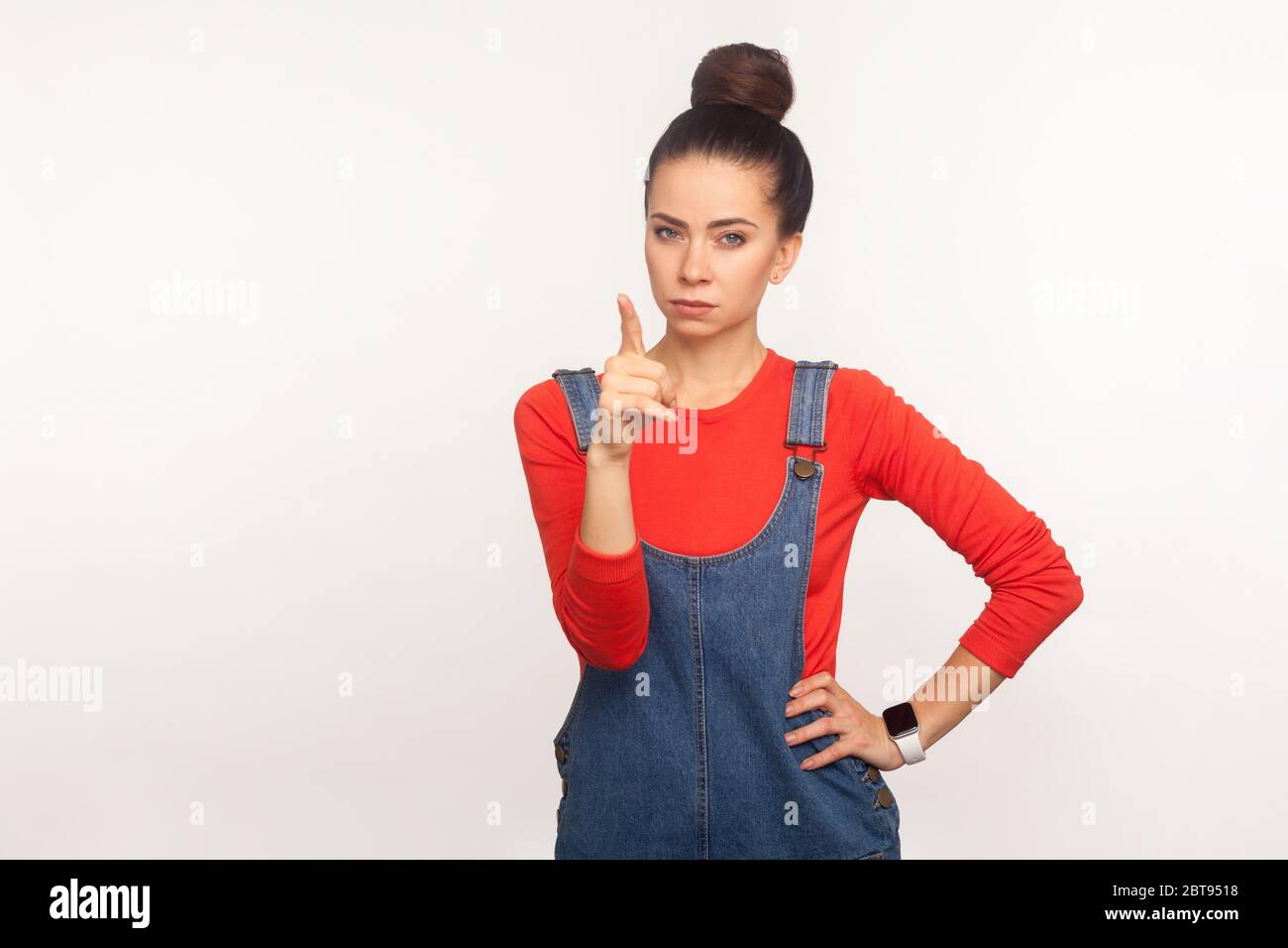 Hé, vous ! Portrait d'une fille en colère bossy avec un pain de cheveux dans des combinaisons en denim qui se disseront pour le mauvais comportement avec le geste de doigt d'admonishing, de la formation et de la formation Banque D'Images