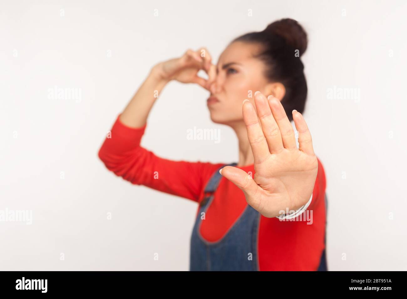 Répulsion à la mauvaise odeur. Portrait d'une fille avec un cheveu dans une combinaison en denim qui retient l'haleine, pinçant son nez et montrant un geste d'arrêt, évitant ainsi les coups de pied Banque D'Images