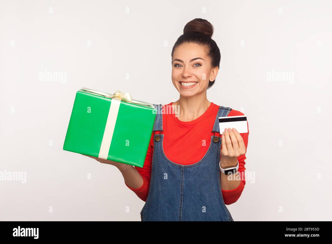 Crédit pour l'achat d'un cadeau de vacances. Portrait d'une fille joyeuse et élégante avec un petit pain en Jean combinaison tenant une boîte cadeau et une carte bancaire, en appréciant les achats Banque D'Images