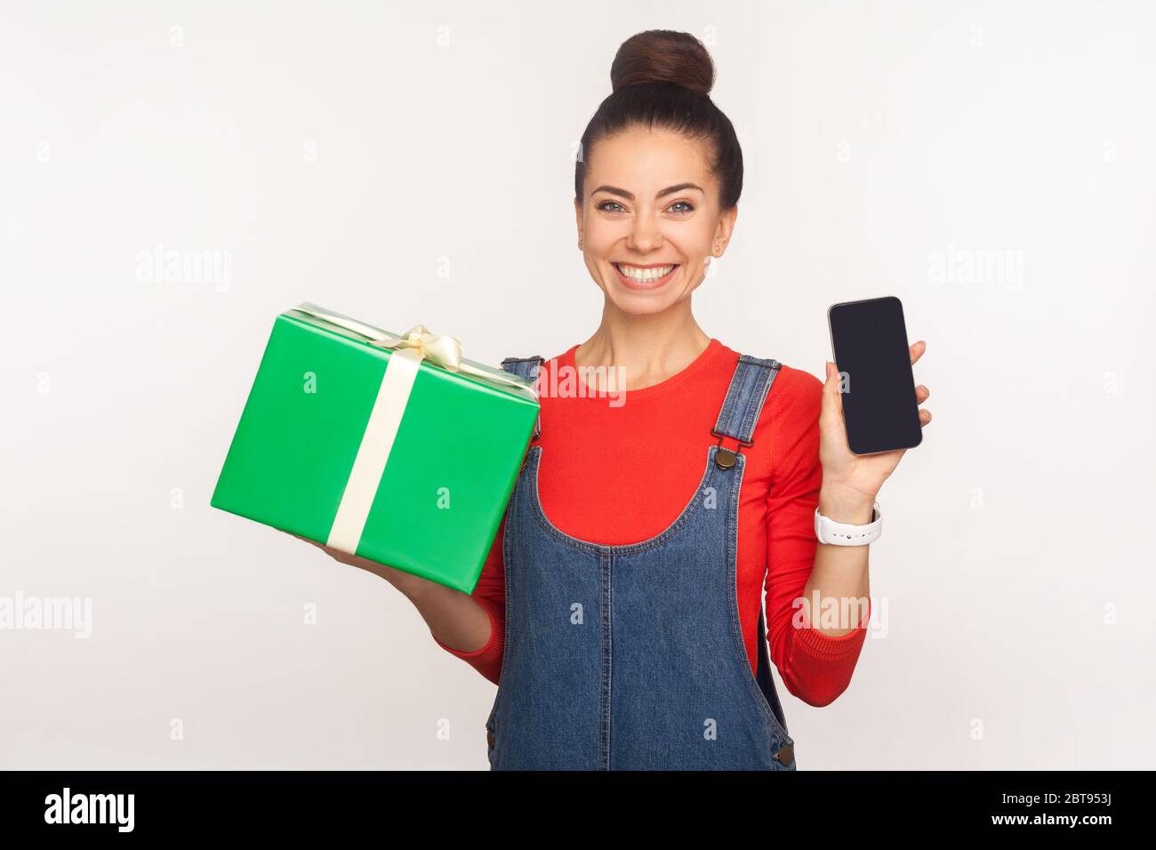 Cadeau pour utilisateur mobile ! Portrait de positive joyeuse jolie fille avec un pain de cheveux dans des combinaisons en denim tenant boîte à présent et téléphone cellulaire, publicité des vacances Banque D'Images