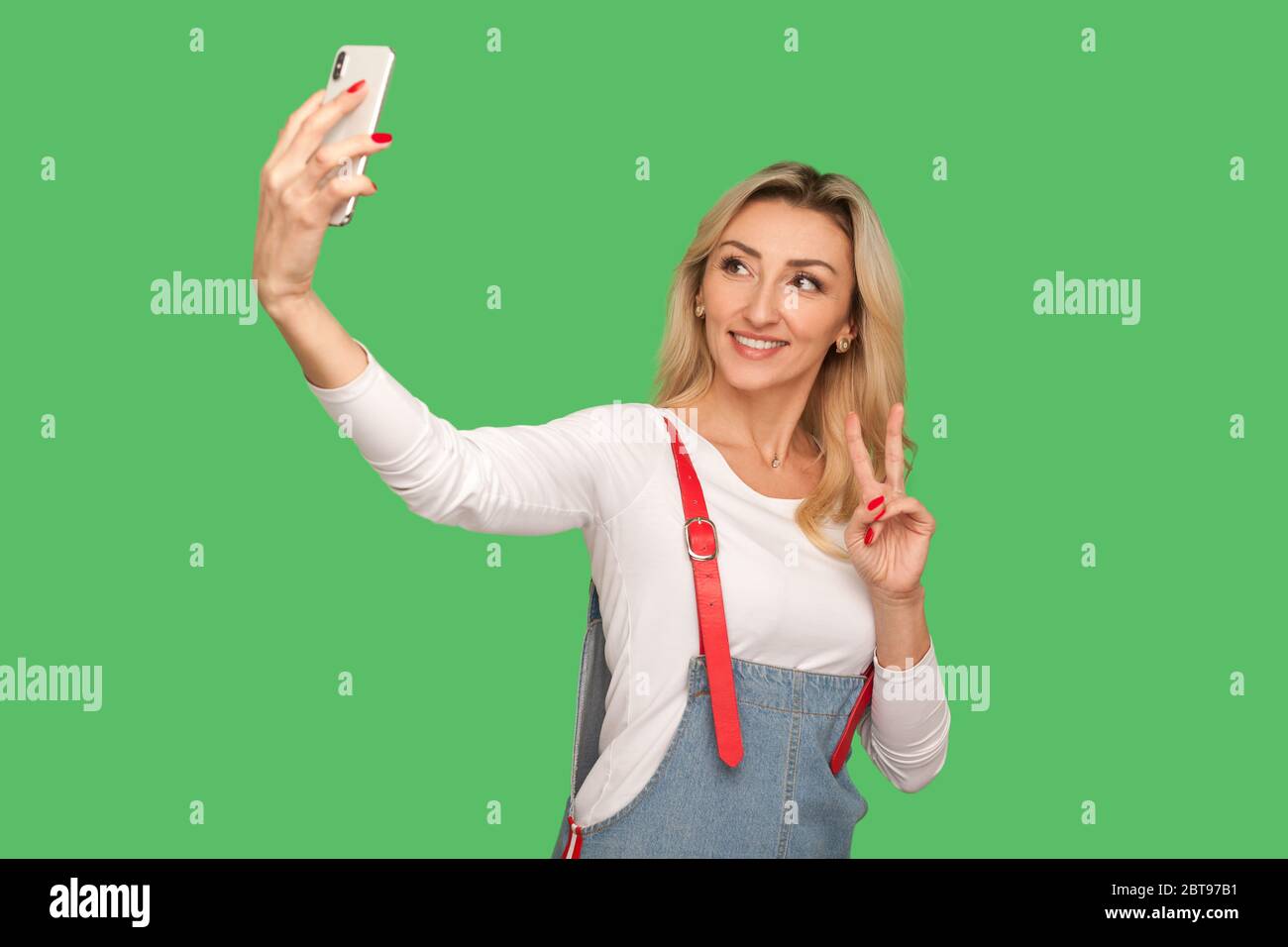 Portrait d'une belle femme adulte avec un sourire amical prenant selfie sur smartphone et montrant le geste de victoire, streaming ou chat sur vidéo c Banque D'Images