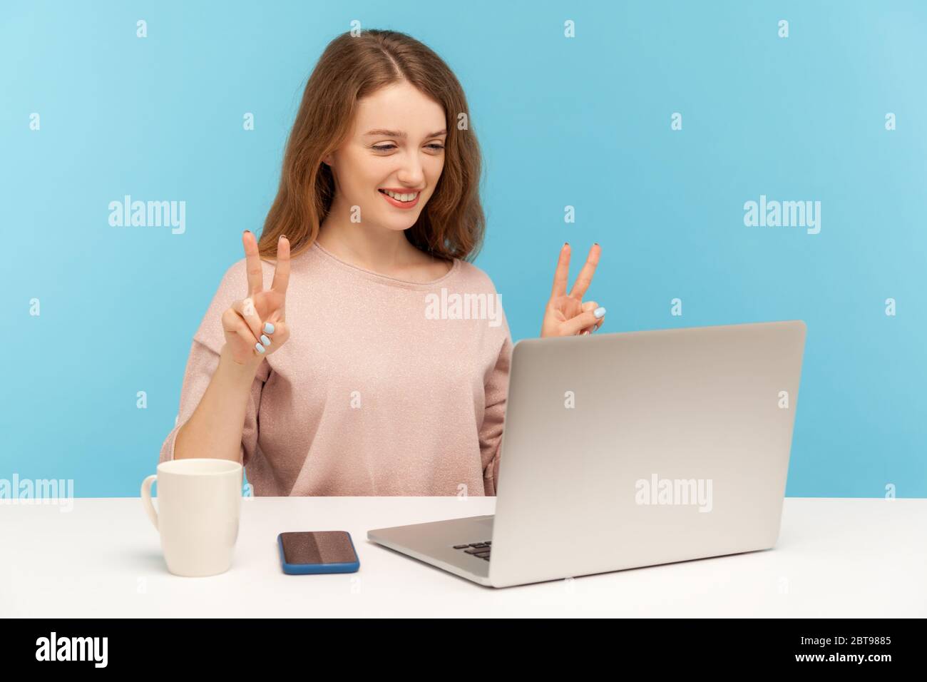 Bonne jeune femme réussie montrant le geste de victoire et souriant sur l'écran d'ordinateur portable, parlant appel vidéo en ligne et de faire le signe v avec deux doigts. Banque D'Images