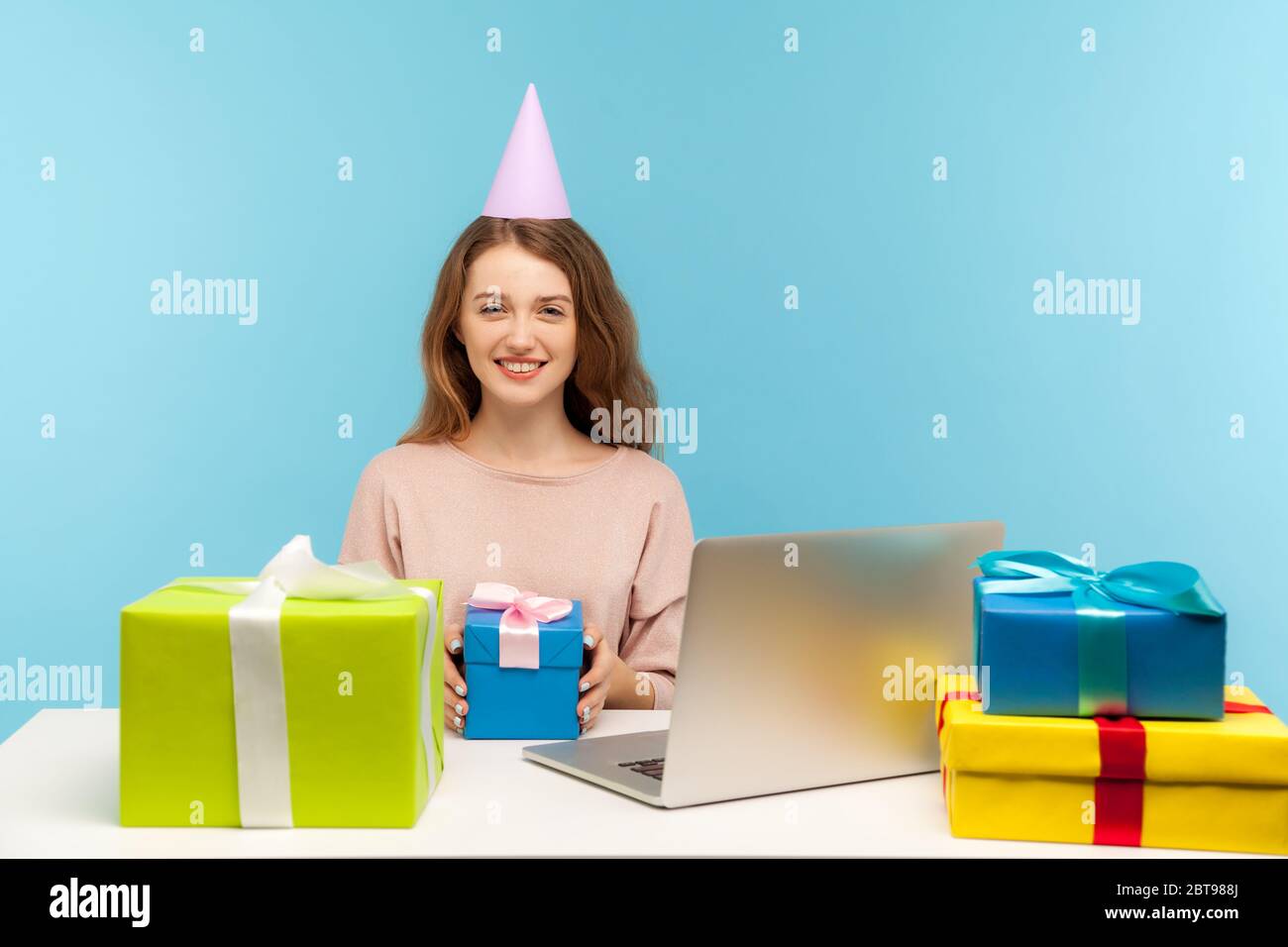 Drôle femme employé de bureau avec le cône de partie assis au lieu de  travail entouré de nombreux cadeaux, souriant amical et tenant boîte cadeau,  célébrant profe Photo Stock - Alamy