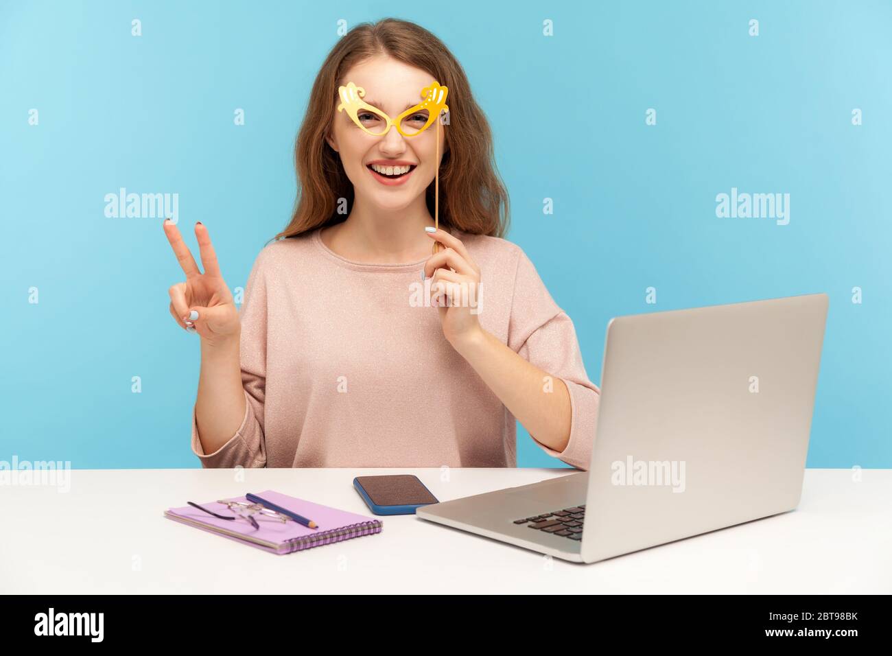 Drôle optimiste femme employé couvrant les yeux avec des lunettes de papier brillant, montrant le geste de victoire et portant l'accessoire de mascarade humoristique, s'amuser Banque D'Images