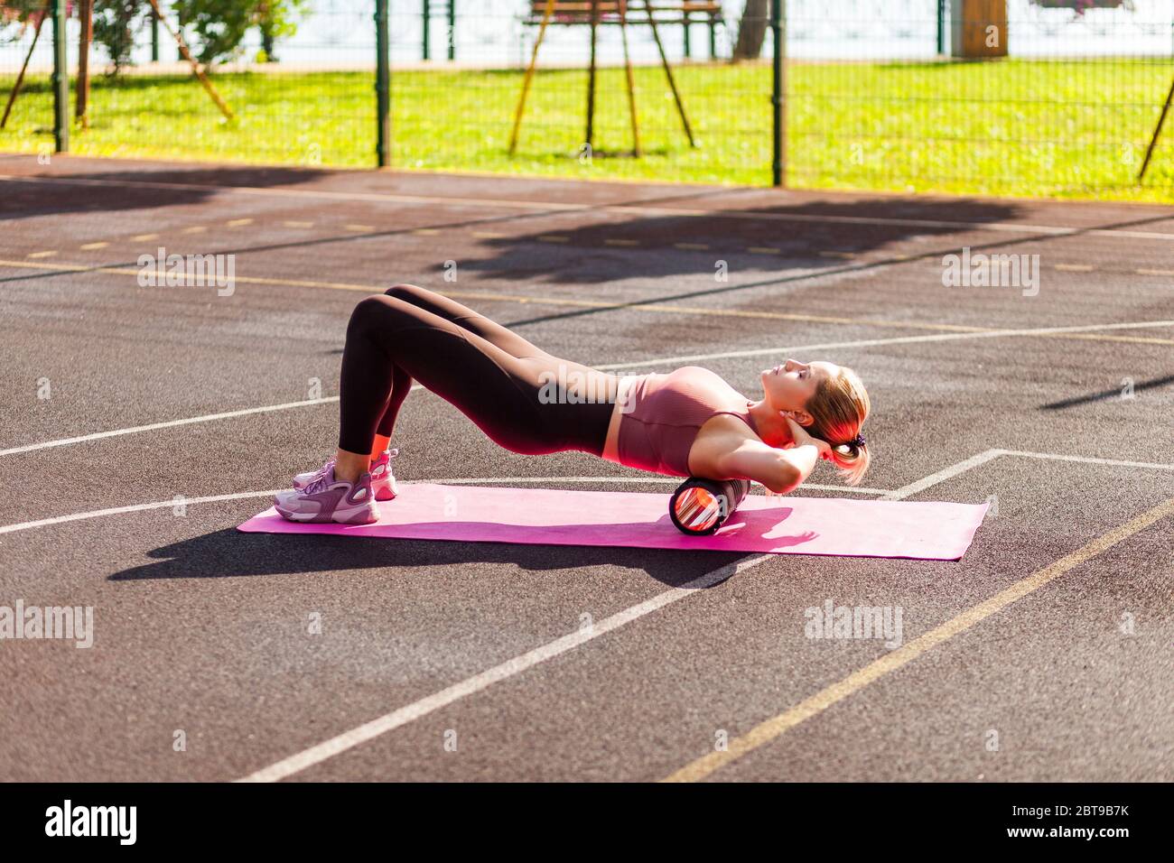Femme blonde sportive s'entraîner sur tapis en extérieur jour d'été, avec un masseur à rouleau en mousse sur le haut du dos pour la détente, l'étirement des muscles de la colonne vertébrale, faire fa Banque D'Images