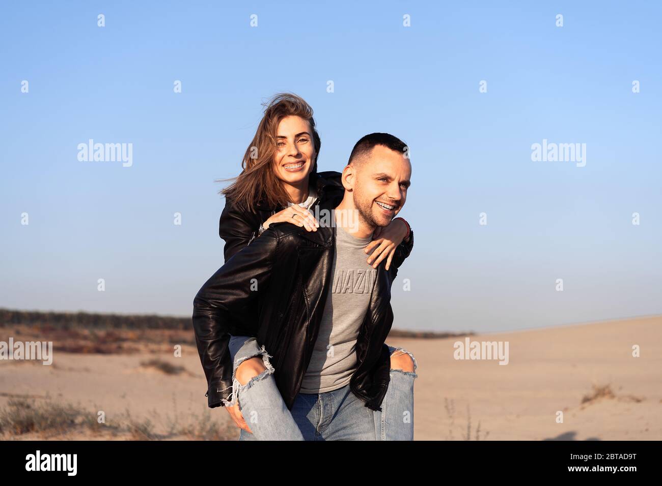 Joyeux et mignon adorable adulte couple veste en cuir et jeans homme avec femme petite amie sur le dos piggy, jouer, rire, sourire et sauter sur le coucher du soleil A. Banque D'Images