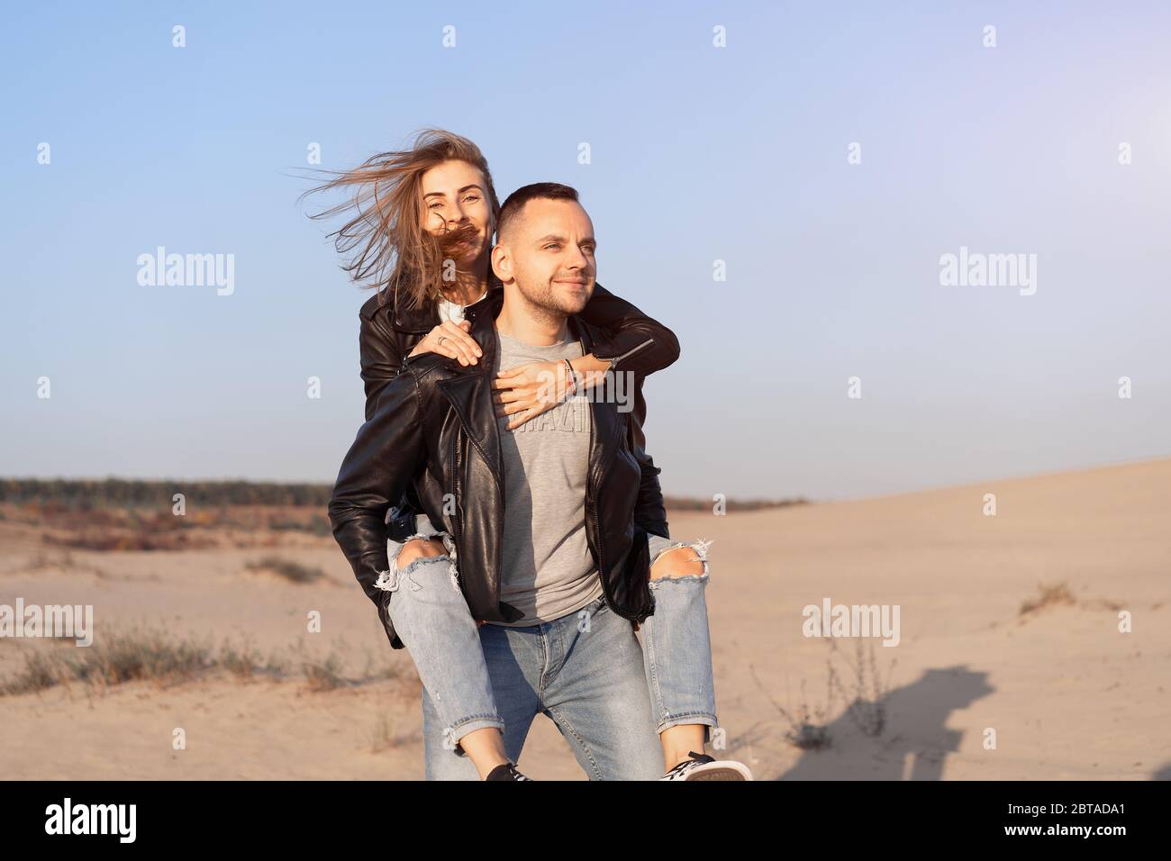 Joyeux et mignon adorable adulte couple veste en cuir et jeans homme avec femme petite amie sur le dos piggy, jouer, rire, sourire et sauter sur le coucher du soleil A. Banque D'Images