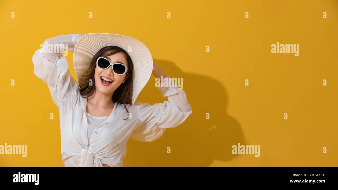 Portrait asiatique belle jeune femme heureuse avec des lunettes de soleil et chapeau souriant gai en été et regardant l'appareil photo isolé sur le studio jaune backgrou Banque D'Images