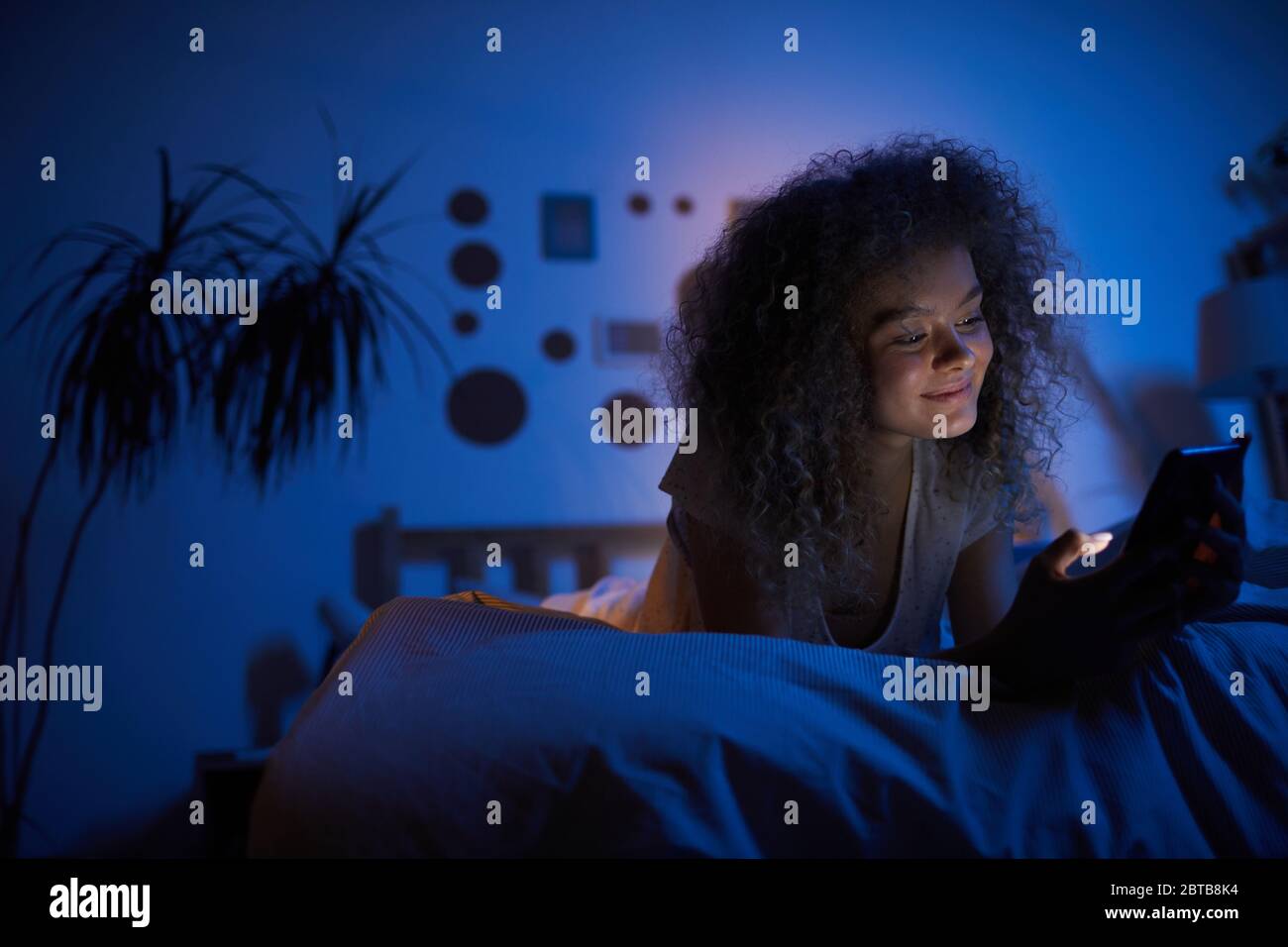 Portrait de jeune femme mauriquement utilisant un smartphone et souriant avec joie en étant allongé sur le lit dans une pièce sombre, espace de copie Banque D'Images