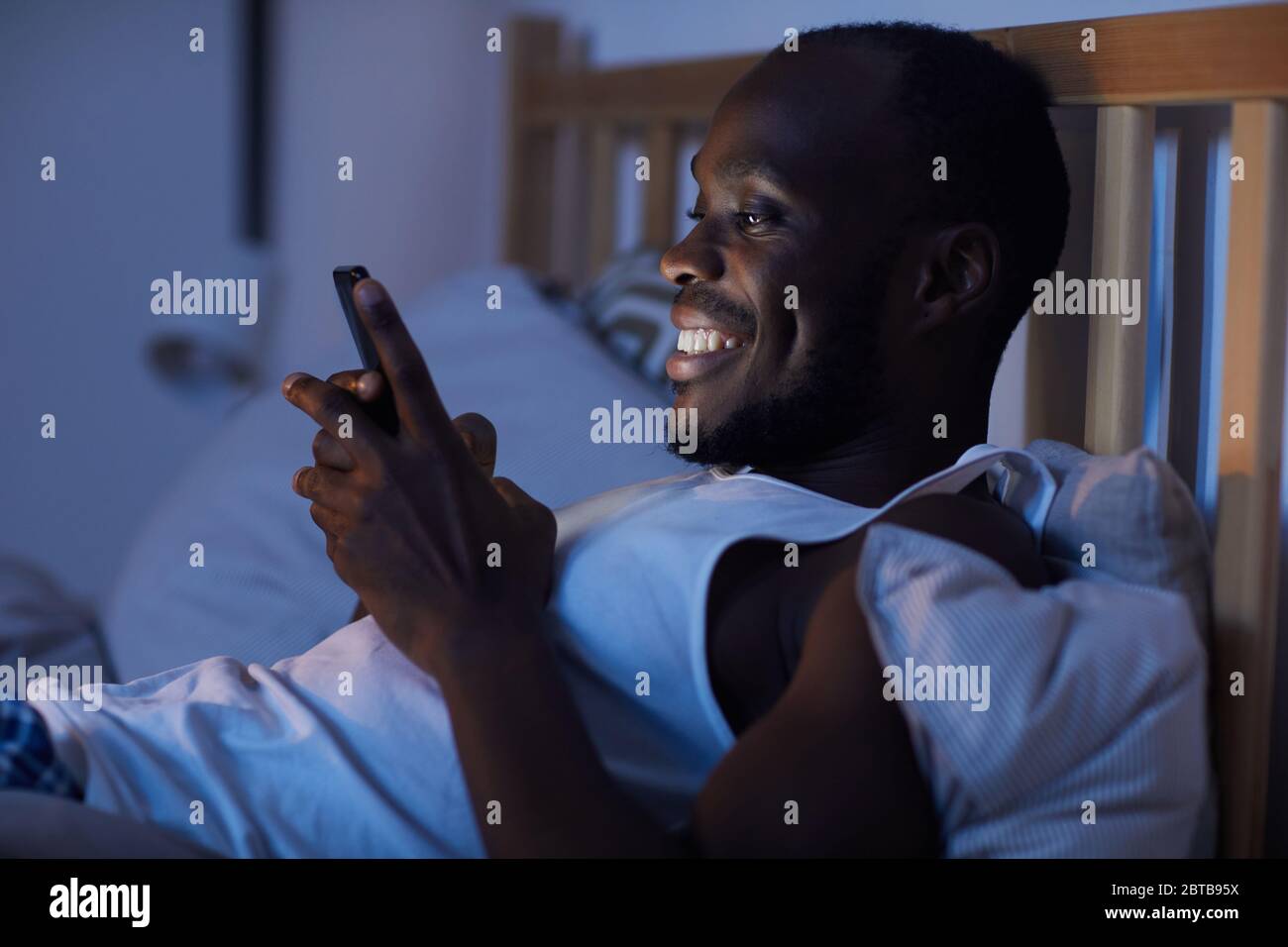 Portrait de l'homme afro-américain souriant en vue latérale, avec un smartphone dans le lit la nuit, espace de copie Banque D'Images