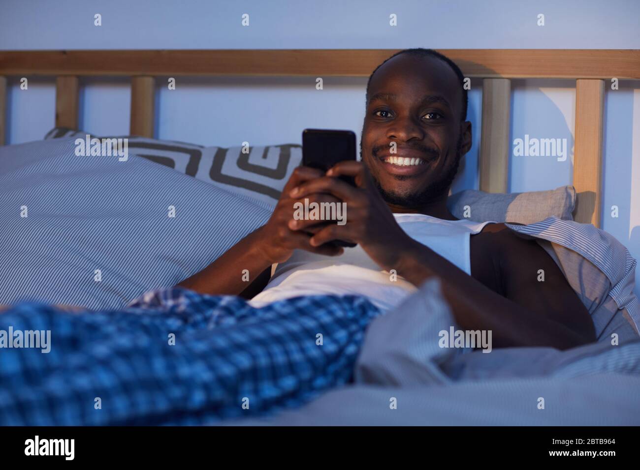 Portrait d'un homme afro-américain souriant utilisant un smartphone dans le lit la nuit et regardant l'appareil photo, l'espace de copie Banque D'Images