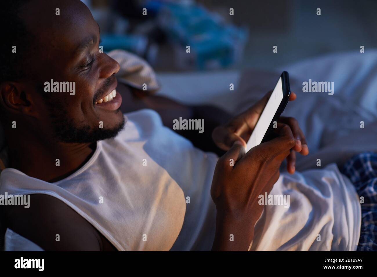 Portrait en grand angle d'un homme afro-américain souriant utilisant un smartphone dans le lit la nuit, espace de copie Banque D'Images