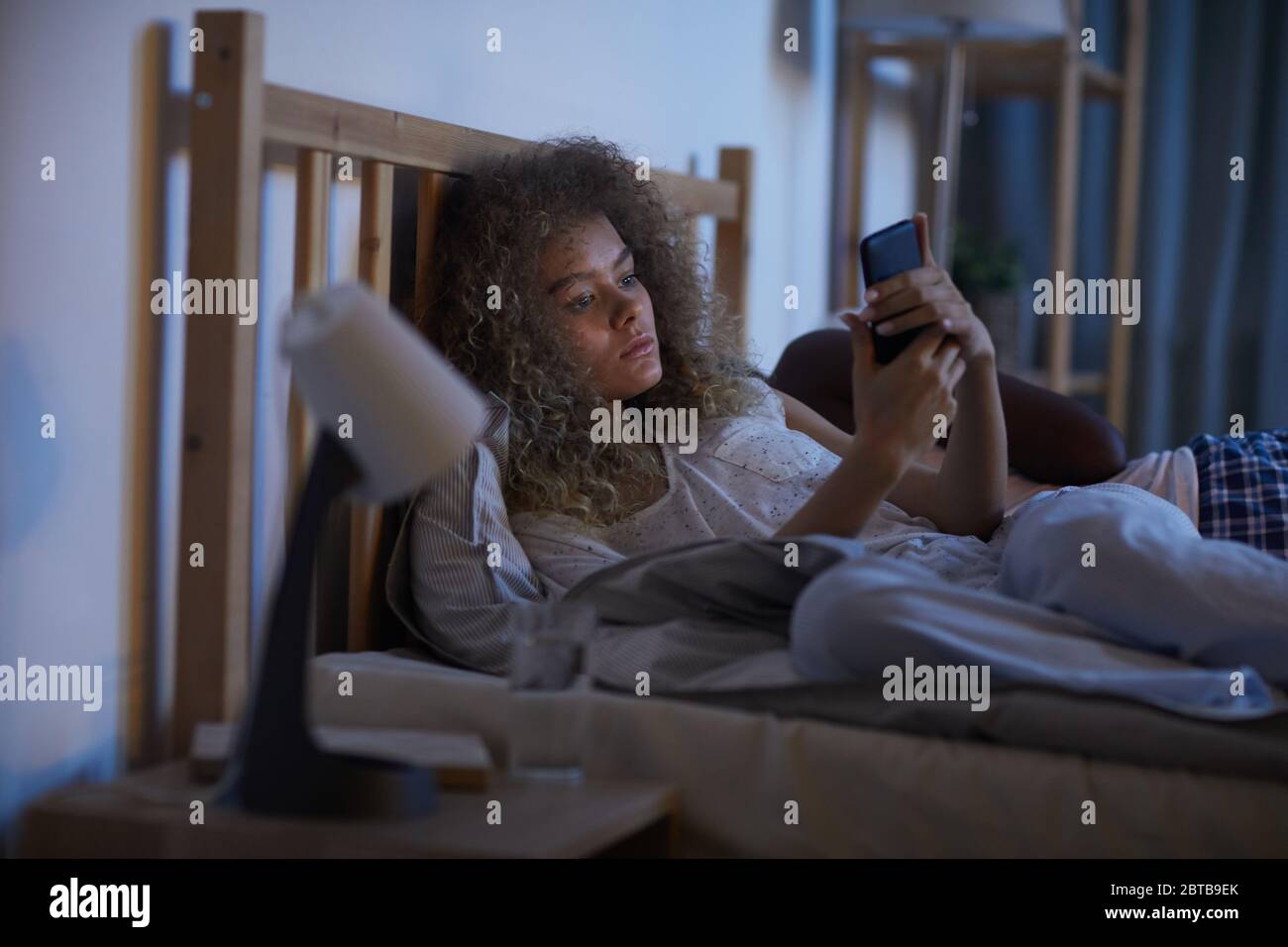 Portrait d'une jeune femme aux cheveux bouclés utilisant un smartphone dans un lit la nuit tout en souffrant d'insomnie, espace de copie Banque D'Images