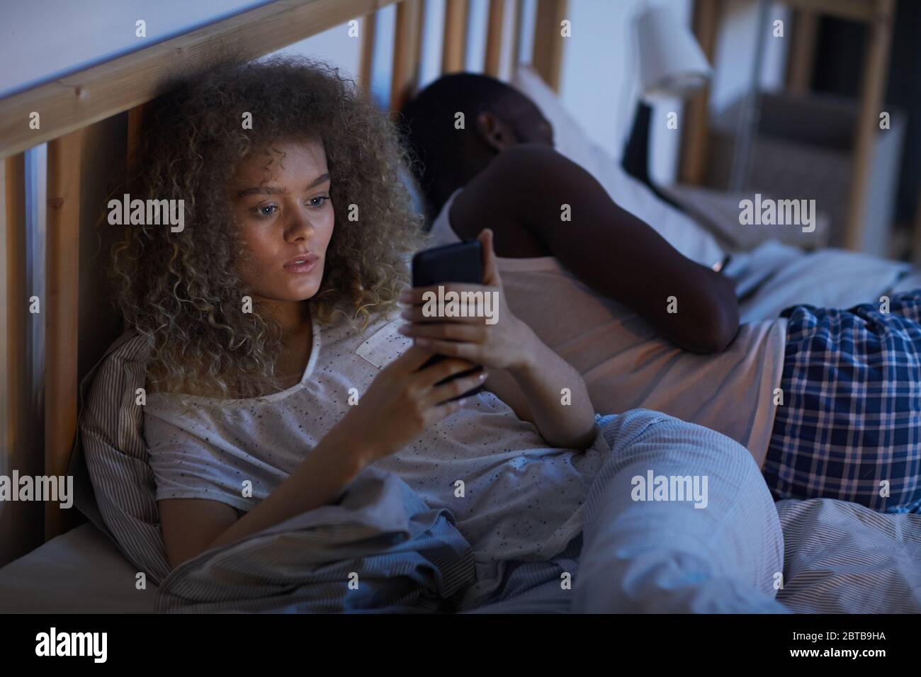 Portrait grand angle de la jeune femme à cheveux bouclés utilisant un smartphone dans le lit la nuit tout en souffrant d'insomnie, espace de copie Banque D'Images