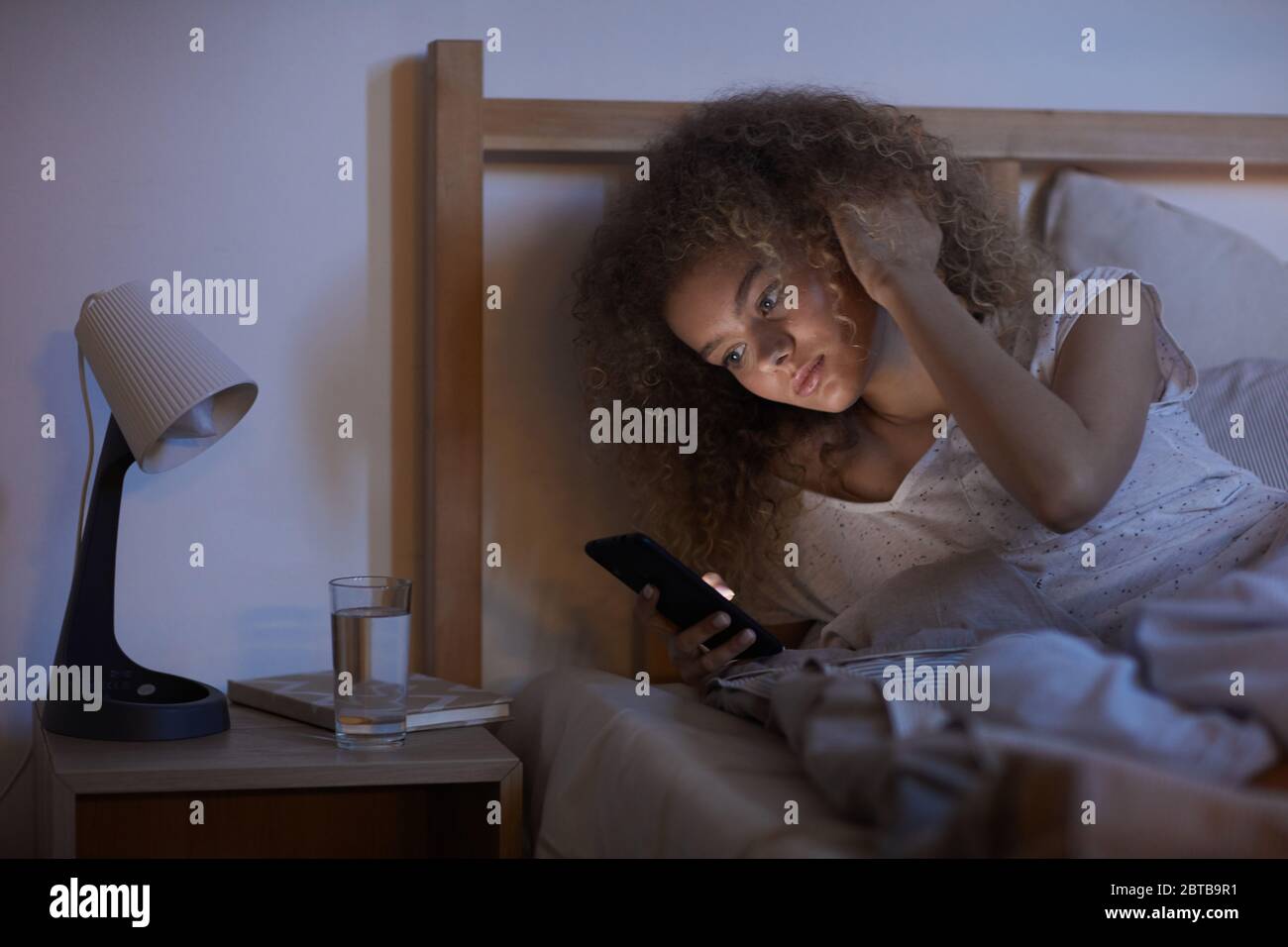 Portrait d'une jeune femme aux cheveux bouclés utilisant un smartphone au lit la nuit, tout en envoyant des SMS ou en naviguant sur Internet, dans un espace de copie Banque D'Images