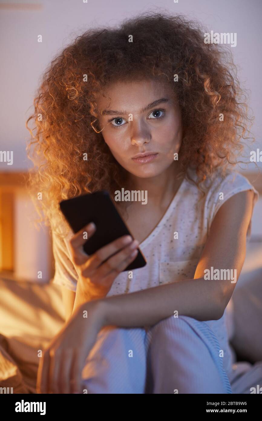 Portrait vertical d'une jeune femme aux cheveux bouclés utilisant un smartphone, assise sur le lit la nuit et regardant l'appareil photo Banque D'Images