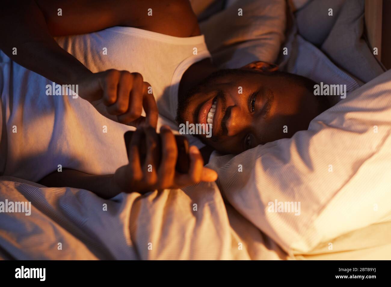 Portrait en grand angle d'un homme afro-américain souriant utilisant un smartphone dans le lit la nuit, tout en envoyant un SMS à une petite amie, dans un espace de copie Banque D'Images