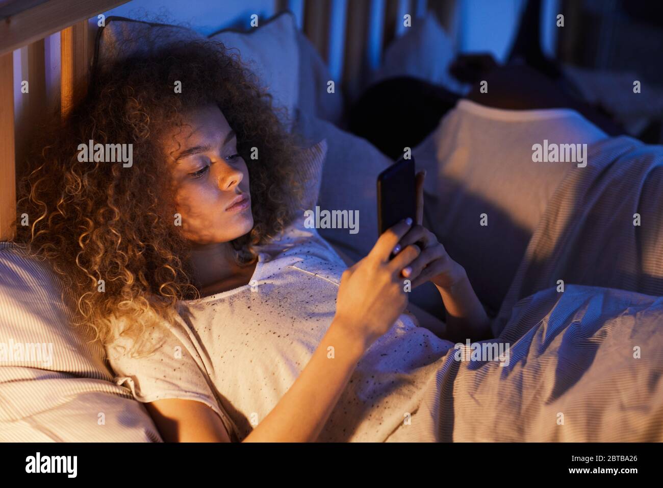 Portrait grand angle de la jeune femme aux cheveux bouclés utilisant un smartphone dans le lit la nuit, en étant couché à côté de son mari ou de son petit ami, espace de copie Banque D'Images
