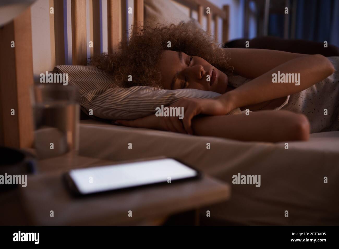 Portrait d'une jeune femme aux cheveux bouclés dormant calmement dans un lit la nuit avec un smartphone éclairé sur une table de nuit, espace de copie Banque D'Images
