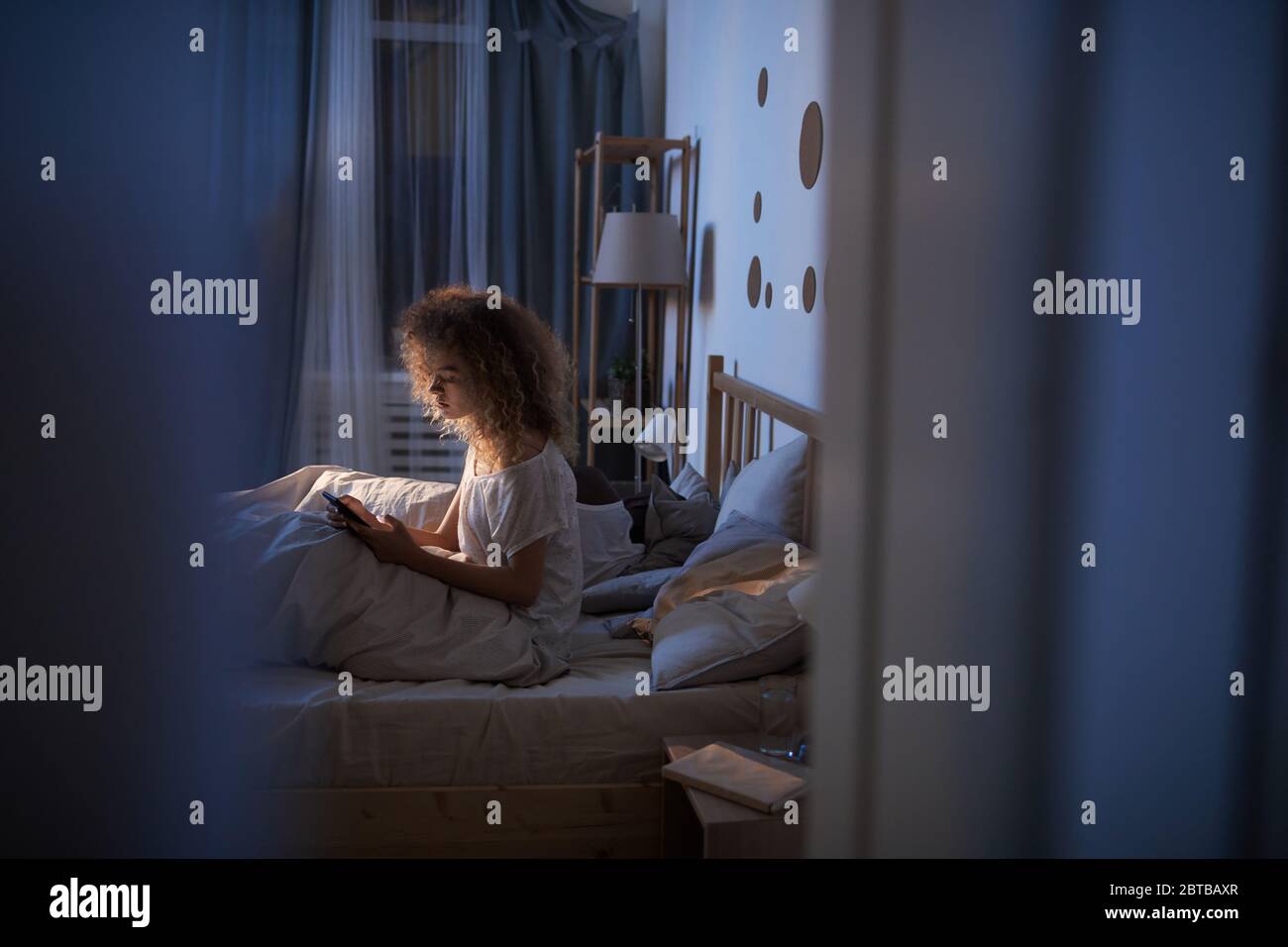 Portrait de la jeune femme aux cheveux bouclés avec une tablette numérique, assise au lit la nuit et Internet, prise de vue par fenêtre, station santé Banque D'Images