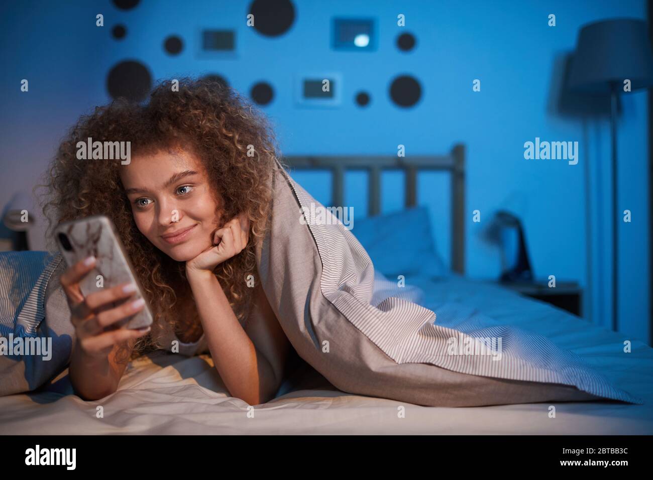 Portrait d'une jeune femme aux cheveux bouclés regardant l'écran du smartphone et souriant, couchée au lit la nuit, espace de copie Banque D'Images